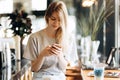 A cute thin blonde girl,dressed in casual style,drinks coffee and looks at her phone in a coffee shop.