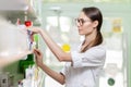 A cute thin lady with dark hair and glasses,wearing a lab coat,takes something from the shelf in an up-to-date pharmacy