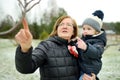 Cute toddler boy and his grandma having fun on a walk in snow covered pine forest on chilly winter day