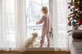 Cute toddler boy in pajama, standing in front of a big french windows with his pet dog, enjoying the snow Royalty Free Stock Photo