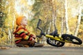 Cute toddler boy sitting on empty road in autumn park looking on falling orange leaves. Child`s bicycle lies nearby. Fall vibes Royalty Free Stock Photo