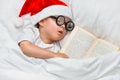 A cute toddler boy is sleeping in a santa hat and round glasses with an old book under a white blanket. Royalty Free Stock Photo