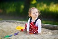 Cute toddler girl playing in sand on outdoor playground. Beautiful baby in red gum trousers having fun on sunny warm Royalty Free Stock Photo