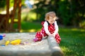 Cute toddler girl playing in sand on outdoor playground. Beautiful baby in red gum trousers having fun on sunny warm Royalty Free Stock Photo