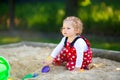 Cute toddler girl playing in sand on outdoor playground. Beautiful baby in red gum trousers having fun on sunny warm Royalty Free Stock Photo