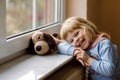 Cute toddler girl sitting by window and looking out. Dreaming child with soft dog toy feeling happy. Lonely kid. Royalty Free Stock Photo