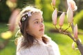 Cute tween girl with with handmade wreath on head with funny face expression looking surprisingly at magnolia tree buds.