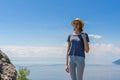 Cute tween tourist girl in hat and backpack standing on cliff top and posing against beautiful landscape of blue sky and Baikal