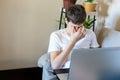 Cute upset boy in white t shirt sitting on the couch in the living room next to laptop and study. Stressed, tired kid. Royalty Free Stock Photo