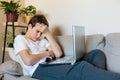 Cute upset boy in white t shirt sitting on the couch in the living room next to laptop and study. Stressed, tired kid. Royalty Free Stock Photo