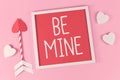 Valentine`s Day composition with picture frame with text `Be Mine`, cupid love arrows, and white hearts on pink background