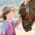 Cute white toddler girl in a rustic style dress caressing brown pony in a field in sunny day Royalty Free Stock Photo