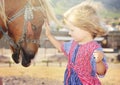 Cute white toddler girl in a rustic style dress caressing brown pony in a field in sunny day Royalty Free Stock Photo