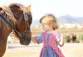 Cute white toddler girl in a rustic style dress caressing brown pony in a field in sunny day Royalty Free Stock Photo