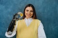 Cute woman with a bionic prosthetic arm holds a gold bitcoin coin and smiles on a blue background.