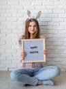 Cute woman in bunny ears holding felt letter board with the words Happy Easter on white brick wall background Royalty Free Stock Photo