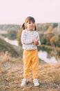 A cute 3-year-old girl with two ponytails and a polka-dot sweater stands on the river bank on a warm autumn day, Royalty Free Stock Photo