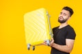 Cute young bearded hipster man holding a heavy yellow suitcase on a yellow background. Concept of heavy load for Royalty Free Stock Photo