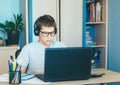 Cute young boy in blue shirt sitting behind desk in his room next to laptop and study. Teenager in earphones makes homework,