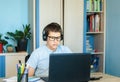 Cute young boy in blue shirt sitting behind desk in his room next to laptop and study. Teenager in earphones makes homework,