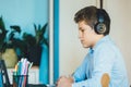 Cute young boy in blue shirt sitting behind desk in his room next to laptop and study. Teenager in earphones makes homework,