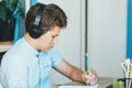 Cute young boy in blue shirt sitting behind desk in his room next to laptop and study. Teenager in earphones makes homework,