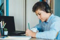 Cute young boy in blue shirt sitting behind desk in his room next to laptop and study. Teenager in earphones makes homework,