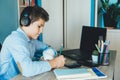 Cute young boy in blue shirt sitting behind desk in his room next to laptop and study. Teenager in earphones makes homework,