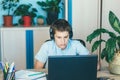 Cute young boy in blue shirt sitting behind desk in his room next to laptop and study. Teenager in earphones makes homework,