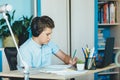 Cute young boy in blue shirt sitting behind desk in his room next to laptop and study. Teenager in earphones makes homework,