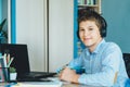 Cute young boy in blue shirt sitting behind desk in his room next to laptop and study. Teenager in earphones makes homework,