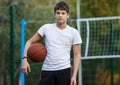 Cute young boy plays basketball on street playground in summer. Teenager in white t-shirt with orange basketball ball outside. Royalty Free Stock Photo