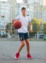 A cute young boy plays basketball on the street playground in summer. Teenager in a white t-shirt with orange basketball ball