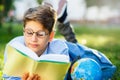 Cute, young boy in round glasses and blue shirt reads book lying on the grass in the park. Education, back to school Royalty Free Stock Photo