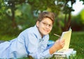 Cute, young boy in round glasses and blue shirt reads book lying on the grass in the park. Education, back to school Royalty Free Stock Photo