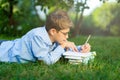 Cute, young boy in round glasses and blue shirt reads book lying on the grass in the park. Education, back to school Royalty Free Stock Photo