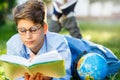 Cute, young boy in round glasses and blue shirt reads book lying on the grass in the park. Education, back to school Royalty Free Stock Photo