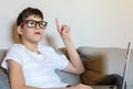 Cute young boy white t shirt sitting on the couch in the living room with his finger up next to laptop and study. Home education, Royalty Free Stock Photo