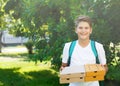 Cute, young boy in white t shirt stands on the grass and holds boxes with pizza in the summer park. Boy eats pizza Royalty Free Stock Photo