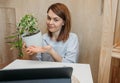 Cute young brunette woman in simple clothes shows SchefflerÃÂ° flower in white pot in her laptop. Online consultation about care of