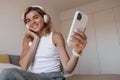 Cute young caucasian woman listening music and holds smartphone sitting on bed in bedroom. Royalty Free Stock Photo