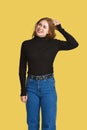 Cute young Caucasian woman with a puzzled and funny expression stands scratching the back of her head. Yellow background Royalty Free Stock Photo