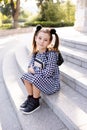 Cute young child girl 5-6 year old wear checkered black and white dress and backpack holding books sitting on stairs outdoors clos Royalty Free Stock Photo