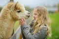 Cute Young Girl Stroking An Alpaca At A Farm Zoo On Autumn Day. Child Feeding A Llama On An Animal Farm. Kid At A Petting Zoo At
