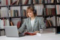 Cute young girl student working on a University project in the library on a laptop, with a book and headphones. Royalty Free Stock Photo