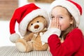 Cute young girl wearing santa hat whispering a secret to her teddy bear christmas present toy. Cheeky kid with teddy bear.