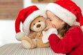 Cute young girl wearing santa hat whispering a secret to her teddy bear christmas present toy. Kid sharing secrets with teddy bear Royalty Free Stock Photo