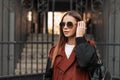 Cute young hipster woman in fashion sunglasses in stylish trench coat with leather black backpack straightens hair near old iron Royalty Free Stock Photo