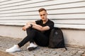 Cute young man in a black T-shirt in stylish black jeans in trendy sneakers with a black sports backpack is resting sitting on the Royalty Free Stock Photo