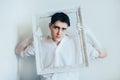 A cute young man wearing a white shirt, holding a blank white frame for a painting. The role of the artist in creating something